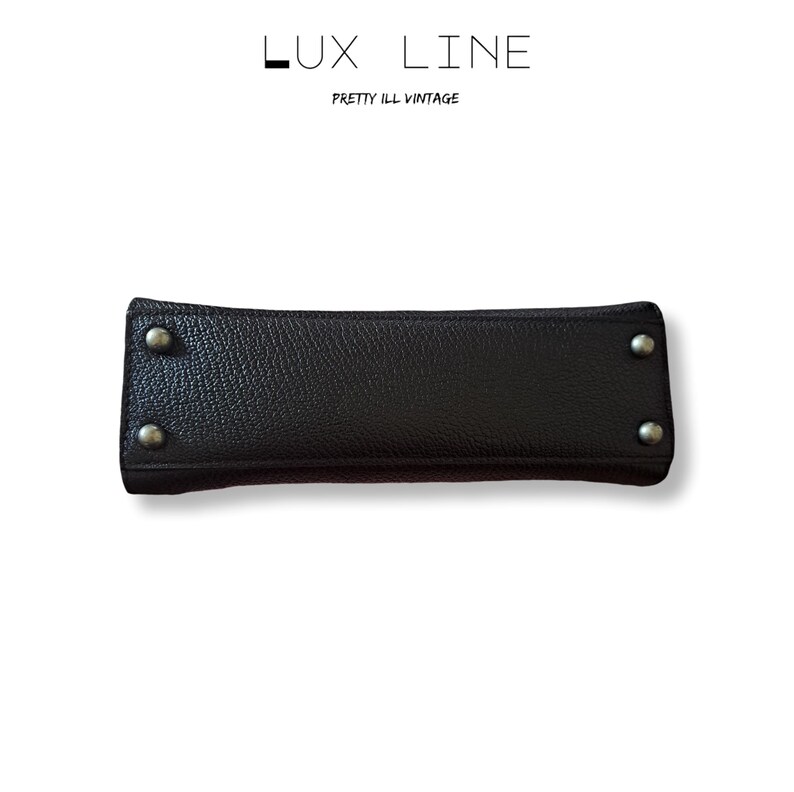 Puede incluir: Un asa de bolso de cuero negro con remaches plateados en cada extremo. El asa es rectangular con una superficie texturizada. El texto "LUX LINE" y "PRETTY ILL VINTAGE" est&aacute; impreso encima.