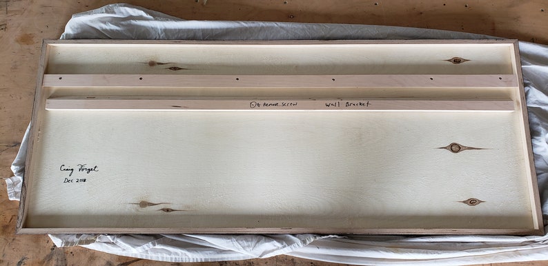 May include: The back of a wooden framed artwork with two wooden strips attached to the back for hanging. The artwork is signed "Craig Forget" and dated "Dec 2018". The wooden strips are labeled "Wall Bracket" and "1/4" Round Stick".