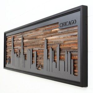 Peut inclure: Une &oelig;uvre d'art murale en bois encadr&eacute;e de noir repr&eacute;sentant une silhouette de l'horizon de Chicago. Le bois est dispos&eacute; en un motif horizontal et l'horizon est d&eacute;coup&eacute; dans le bois. Le cadre est noir et le bois est de diff&eacute;rentes nuances de brun. Le mot "CHICAGO" est d&eacute;coup&eacute; dans le bois en haut de la pi&egrave;ce.