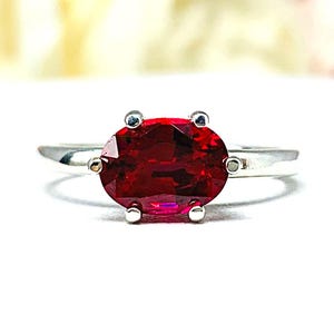 May include: A silver ring featuring a large, oval-cut, deep red gemstone. The gemstone is set with four small prongs. The ring is displayed on a white surface, with blurred floral elements in the background.