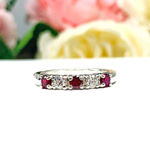May include: A silver-toned ring featuring an alternating row of three round, deep red stones and two clear, sparkling stones set in a prong setting. The band is thin and metallic, showcasing a symmetrical design with five stones in total.