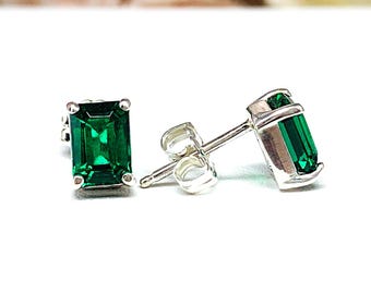 May include: A pair of emerald-cut green gemstone stud earrings set in silver metal. Each earring features a rectangular green stone held by four prongs, with a silver post and butterfly clutch backing visible on one of the earrings.