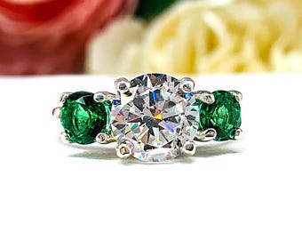 May include: A silver ring with a large, round, clear center stone flanked by two smaller, round, green stones. The ring is set against a blurred background of pink and yellow flowers.