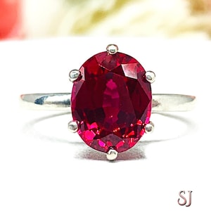 May include: A silver ring featuring a large, oval-cut, deep red gemstone. The gemstone is set with six prongs. The ring is displayed against a white background with blurred pink and white flowers in the background.
