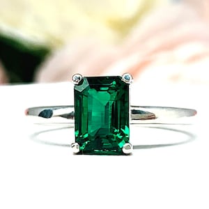 May include: A silver ring featuring a rectangular, emerald-cut green gemstone. The ring has a simple band and is set against a soft, blurred floral background. The initials "SJ" are visible in the lower right corner.