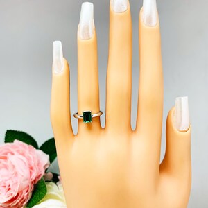 May include: A silver ring with a rectangular emerald-cut green gemstone. The ring is displayed on a mannequin hand with long, white-tipped nails. The text "7X5MM" is visible in the upper left corner. Pink and white flowers are in the background.