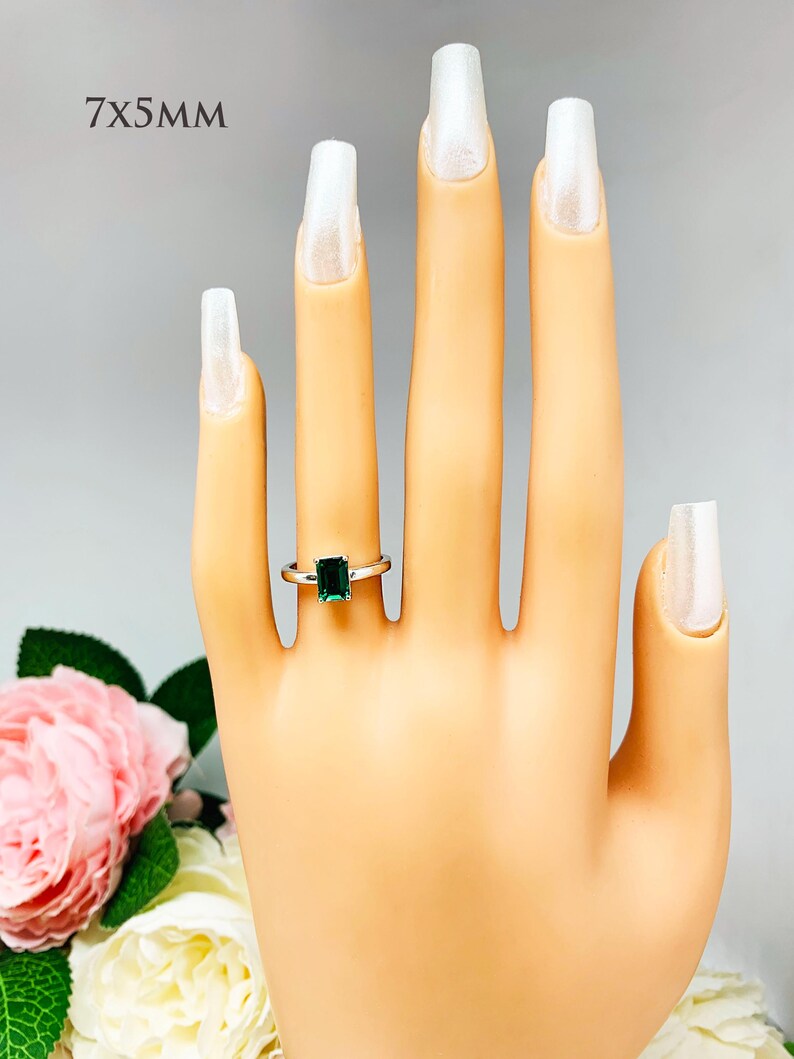 May include: A silver ring with a rectangular emerald-cut green gemstone. The ring is displayed on a mannequin hand with long, white-tipped nails. The text "7X5MM" is visible in the upper left corner. Pink and white flowers are in the background.
