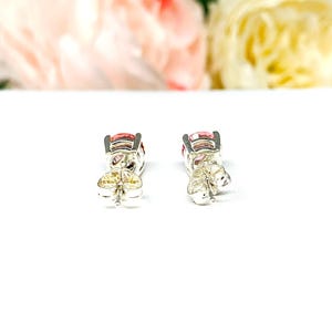 A pair of four prong silver stud earrings set with round lab created padparadscha sapphires. The basket earrings are on a white surface with the both earrings facing toward the back to display the center post and butterfly friction clasps.