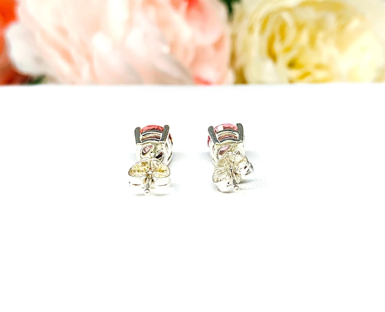 A pair of four prong silver stud earrings set with round lab created padparadscha sapphires. The basket earrings are on a white surface with the both earrings facing toward the back to display the center post and butterfly friction clasps.