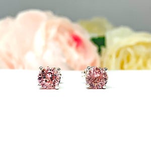 A pair of four prong silver stud earrings set with round lab created padparadscha sapphires. The basket earrings are on a white surface with the both earrings facing front to showcase the mixed brilliant faceting and peachy pink color of the stones.