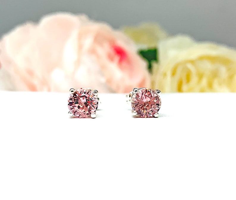 A pair of four prong silver stud earrings set with round lab created padparadscha sapphires. The basket earrings are on a white surface with the both earrings facing front to showcase the mixed brilliant faceting and peachy pink color of the stones.