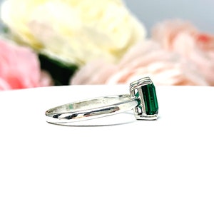 Sterling silver solitaire ring featuring an emerald-cut lab-created emerald. The ring is displayed on a white surface with soft-focus pink and white flowers in the background.