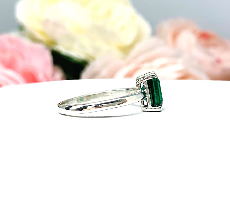 Sterling silver solitaire ring featuring an emerald-cut lab-created emerald. The ring is displayed on a white surface with soft-focus pink and white flowers in the background.
