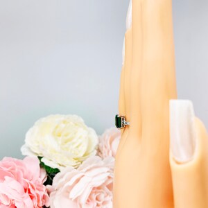 Mannequin hand with long white shimmery nails, displayed against a soft white-gray background with pink and white floral accents. The hand is wearing a sterling silver solitaire ring featuring an 8x6mm emerald-cut lab-created emerald.