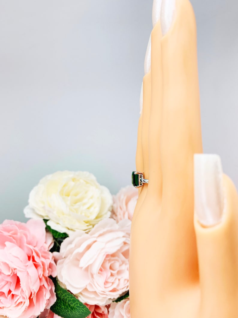 Mannequin hand with long white shimmery nails, displayed against a soft white-gray background with pink and white floral accents. The hand is wearing a sterling silver solitaire ring featuring an 8x6mm emerald-cut lab-created emerald.