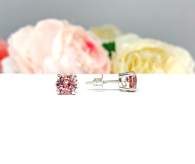 A pair of four prong silver stud earrings set with round lab created padparadscha sapphires. The basket earrings are on a white surface with the left earring facing front to showcase the mixed brilliant faceting and peachy pink color of the stones, and the right earring is facing toward the right to display the center post and butterfly friction clasp.
