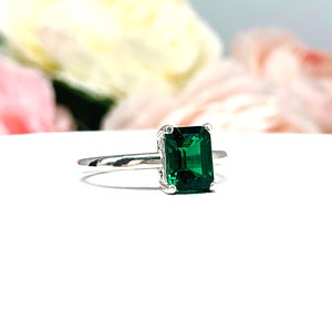 Sterling silver solitaire ring featuring an emerald-cut lab-created emerald. The ring is displayed on a white surface with soft-focus pink and white flowers in the background.