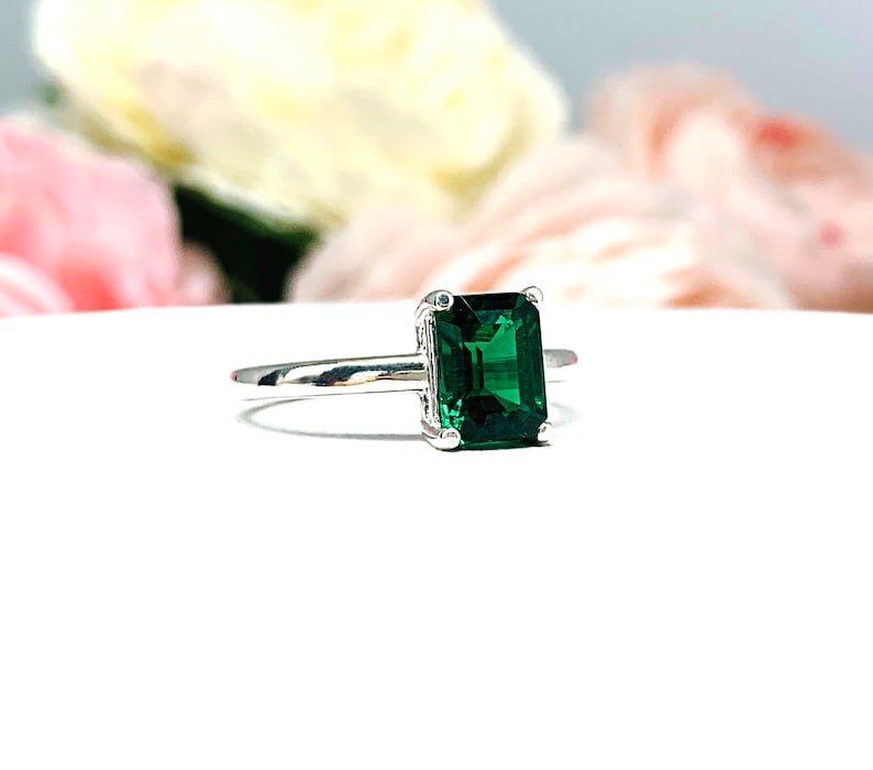 Sterling silver solitaire ring featuring an emerald-cut lab-created emerald. The ring is displayed on a white surface with soft-focus pink and white flowers in the background.