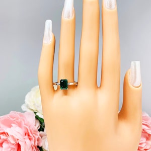 Mannequin hand with long white shimmery nails, displayed against a soft white-gray background with pink and white floral accents. The hand is wearing a sterling silver solitaire ring featuring an 8x6mm emerald-cut lab-created emerald.