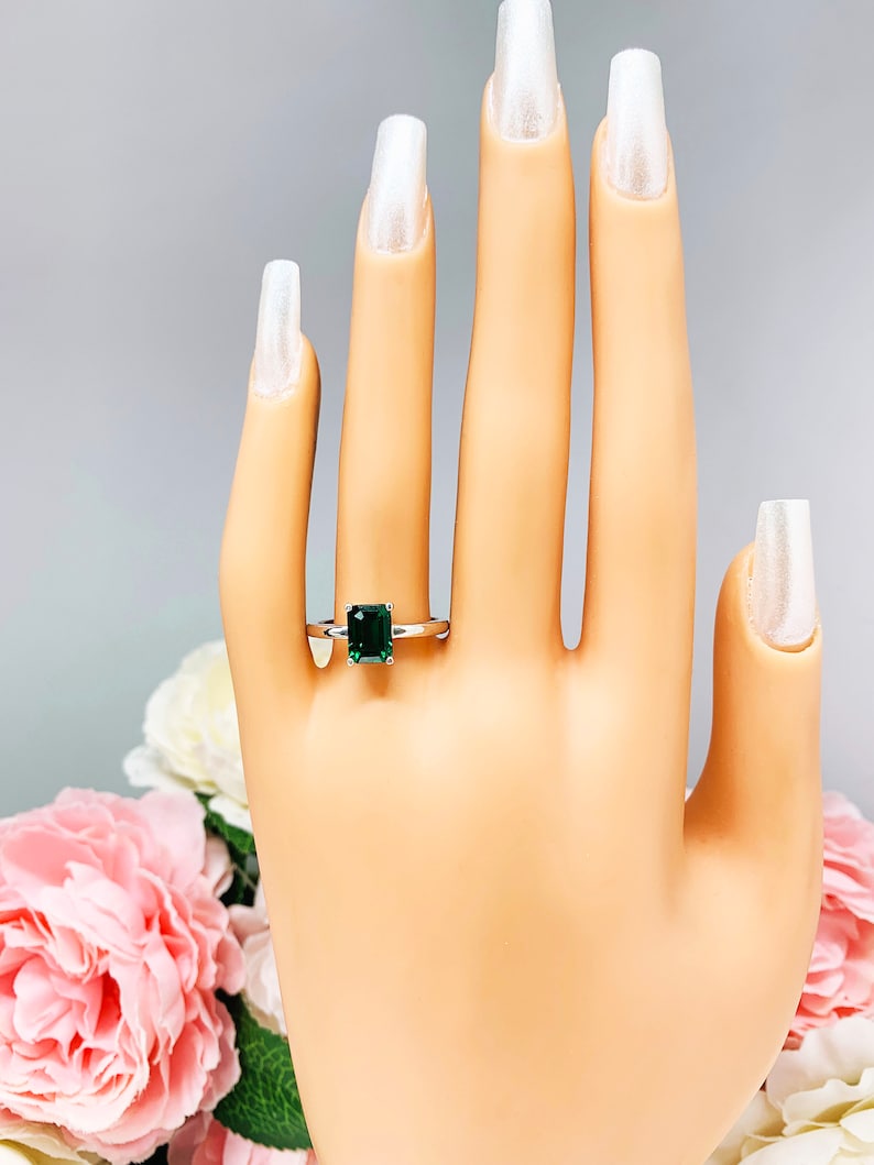 Mannequin hand with long white shimmery nails, displayed against a soft white-gray background with pink and white floral accents. The hand is wearing a sterling silver solitaire ring featuring an 8x6mm emerald-cut lab-created emerald.