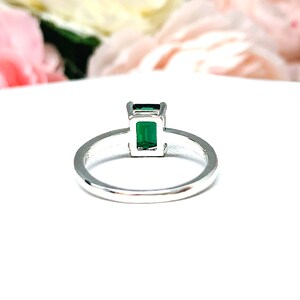 Sterling silver solitaire ring featuring an emerald-cut lab-created emerald. The ring is displayed on a white surface with soft-focus pink and white flowers in the background.