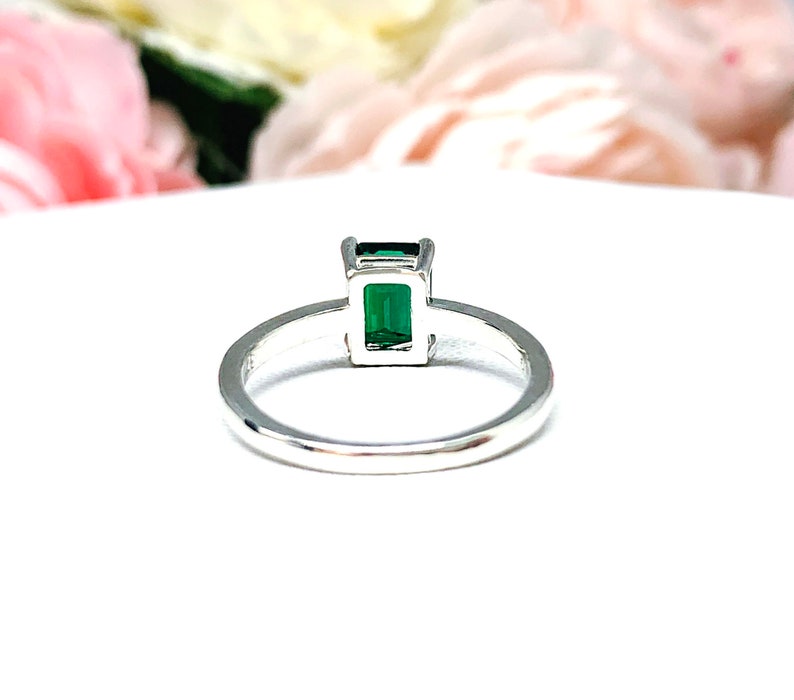 Sterling silver solitaire ring featuring an emerald-cut lab-created emerald. The ring is displayed on a white surface with soft-focus pink and white flowers in the background.