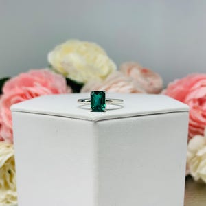 Unedited photo of a sterling silver solitaire ring featuring an emerald-cut lab-created emerald. The ring is displayed on a white surface with soft-focus pink and white flowers in the background.