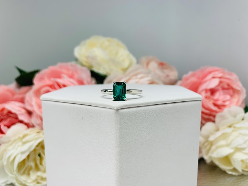 Unedited photo of a sterling silver solitaire ring featuring an emerald-cut lab-created emerald. The ring is displayed on a white surface with soft-focus pink and white flowers in the background.