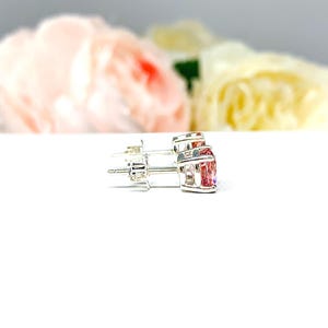 A pair of four prong silver stud earrings set with round lab created padparadscha sapphires. The basket earrings are on a white surface with the both earrings facing toward the right to display the center post and butterfly friction clasps.