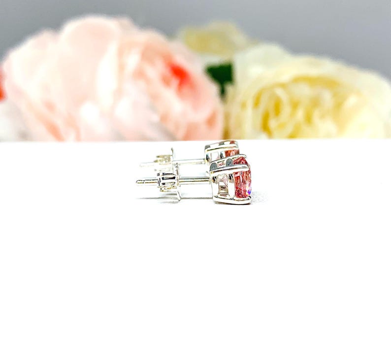A pair of four prong silver stud earrings set with round lab created padparadscha sapphires. The basket earrings are on a white surface with the both earrings facing toward the right to display the center post and butterfly friction clasps.