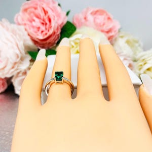 May include: A silver ring with a rectangular, emerald-green gemstone. The ring is displayed on a mannequin hand, with a blurred background of pink and white flowers.