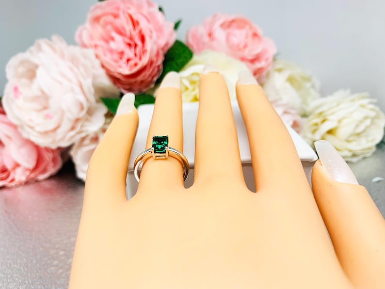 May include: A silver ring with a rectangular, emerald-green gemstone. The ring is displayed on a mannequin hand, with a blurred background of pink and white flowers.