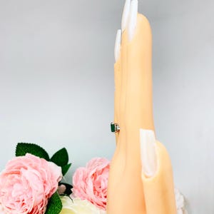 May include: A silver ring with a rectangular emerald-cut green gemstone and small white accent stones. The ring is displayed on a mannequin hand with long, white fingernails. Pink roses and green leaves are in the background.