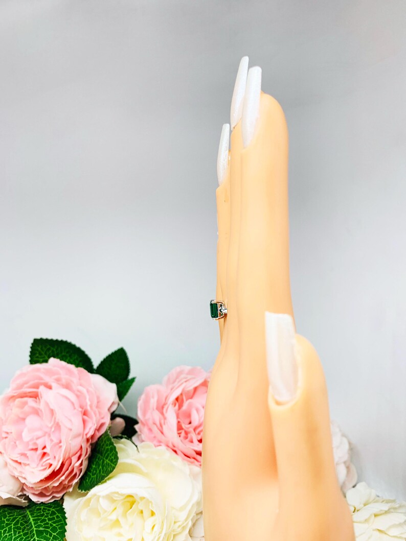 May include: A silver ring with a rectangular emerald-cut green gemstone and small white accent stones. The ring is displayed on a mannequin hand with long, white fingernails. Pink roses and green leaves are in the background.