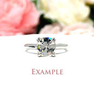 May include: A silver engagement ring with a large, square-cut diamond. The ring is set on a white surface with blurred pink and white flowers in the background. The word "EXAMPLE" is printed in pink.