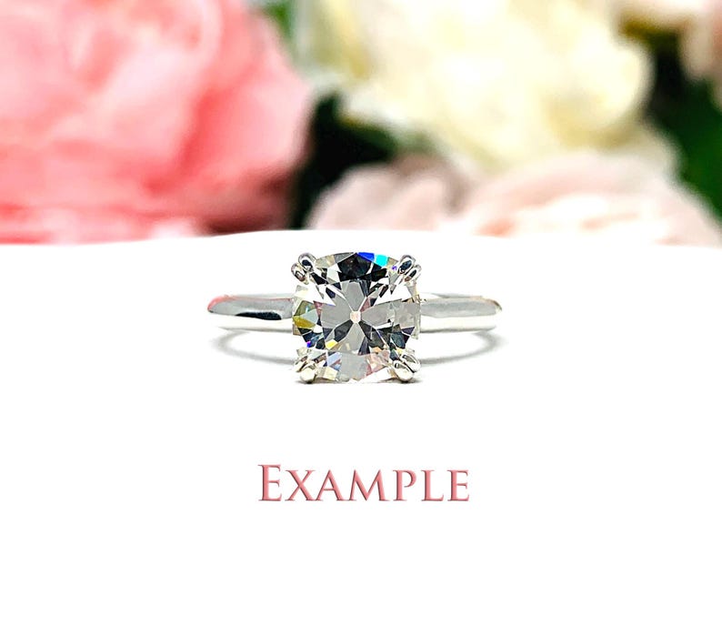 May include: A silver engagement ring with a large, square-cut diamond. The ring is set on a white surface with blurred pink and white flowers in the background. The word "EXAMPLE" is printed in pink.