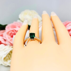 Mannequin hand with long white shimmery nails, displayed against a soft white-gray background with pink and white floral accents. The hand is wearing a sterling silver solitaire ring featuring an 8x6mm emerald-cut lab-created emerald.