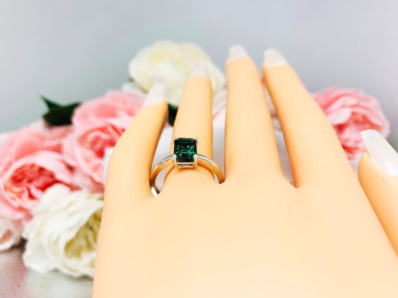 Mannequin hand with long white shimmery nails, displayed against a soft white-gray background with pink and white floral accents. The hand is wearing a sterling silver solitaire ring featuring an 8x6mm emerald-cut lab-created emerald.