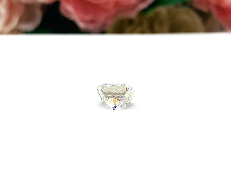 May include: A clear, heart-shaped gemstone with a faceted surface. The gemstone reflects light, displaying a rainbow of colors. The gem is set against a white background, with blurred pink and green floral elements in the background.