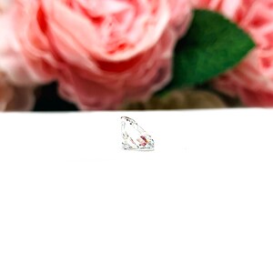 May include: A clear, faceted gemstone with a pear-like shape, resting on a white surface. The gemstone reflects light, showcasing its internal facets. Soft pink roses and green leaves are blurred in the background.