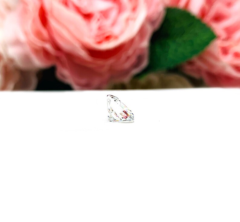 May include: A clear, faceted gemstone with a pear-like shape, resting on a white surface. The gemstone reflects light, showcasing its internal facets. Soft pink roses and green leaves are blurred in the background.