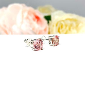 A pair of four prong silver stud earrings set with round lab created padparadscha sapphires. The basket earrings are on a white surface with the both earrings facing slightly toward the right to showcase the mixed brilliant faceting and peachy pink color of the stones and to display the center post and butterfly friction clasps.