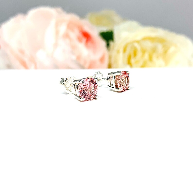 A pair of four prong silver stud earrings set with round lab created padparadscha sapphires. The basket earrings are on a white surface with the both earrings facing slightly toward the right to showcase the mixed brilliant faceting and peachy pink color of the stones and to display the center post and butterfly friction clasps.