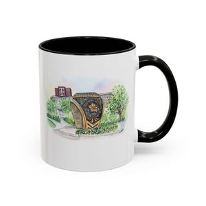 Pode incluir: Caneca de cerâmica branca com borda e alça pretas. A caneca apresenta uma ilustração em aquarela de um monumento de anel dourado com uma estrela e um edifício ao fundo.