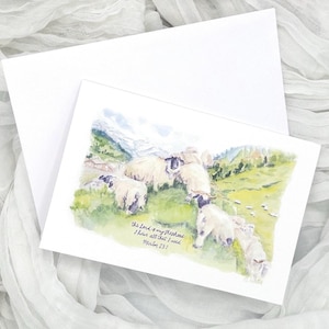 May include: A white greeting card with a watercolor painting of sheep grazing in a green pasture with mountains in the background. The text "The Lord is my shepherd, I have all that I need, Psalm 23:1" is written on the card.