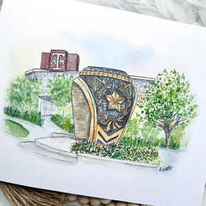 May include: Watercolor painting of a gold and blue ring with a star and a wreath. The ring is in front of a building with the letters "T" and "AM" on it. The painting is on a white background.