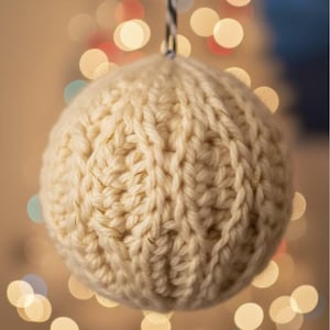May include: A cream-coloured, knitted Christmas bauble with a textured surface, suspended by a black and white string. The background shows blurred, warm-toned lights, creating a festive atmosphere. The bauble is spherical.