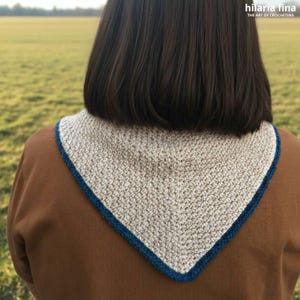 May include: A beige crocheted triangular scarf with a dark blue border. The scarf is worn over a brown sweater. The text "hilaria fina THE ART OF CROCHETING" is in the upper right corner.