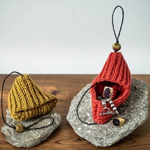 May include: Two handmade knitted pouches in mustard yellow and rust red, each with a black cord and a decorative bead. The red pouch holds jewelry, including a ring and a pendant. The pouches are displayed on gray stones.