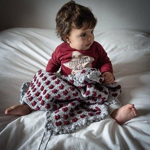 May include: A young child wearing a red shirt with the text "little girl" and a star design is sitting on a white bed with a gray and red crocheted blanket.